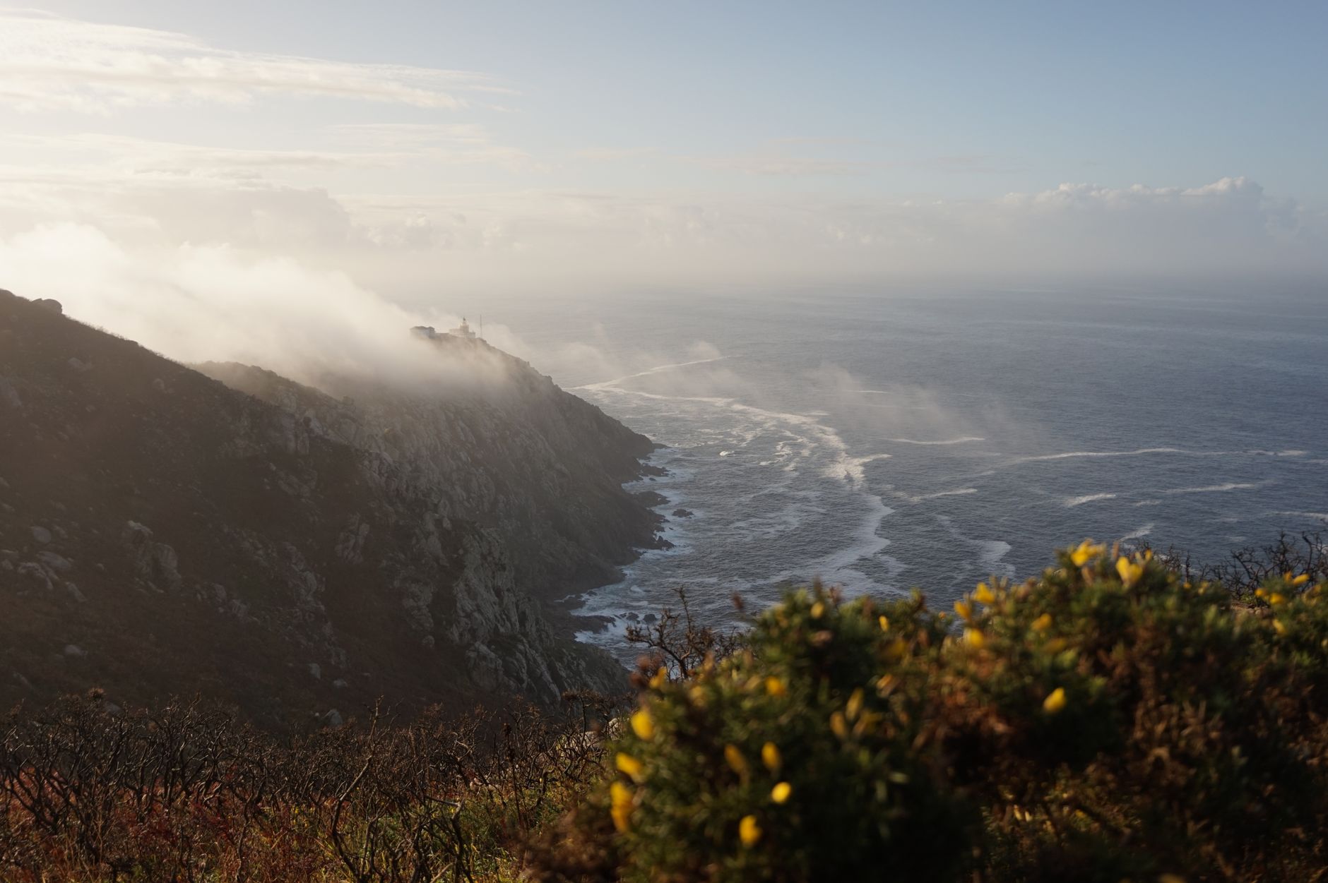 Sunrise view of Fisterra's rugged coast with misty waves and distant lighthouse.