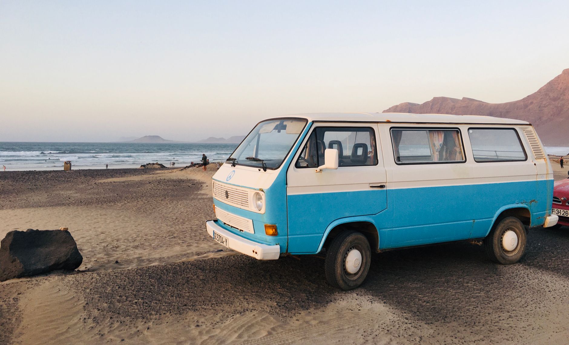 A blue vintage Volkswagen van parked on a sandy beach in Teguise, Spain, with a ocean backdrop.