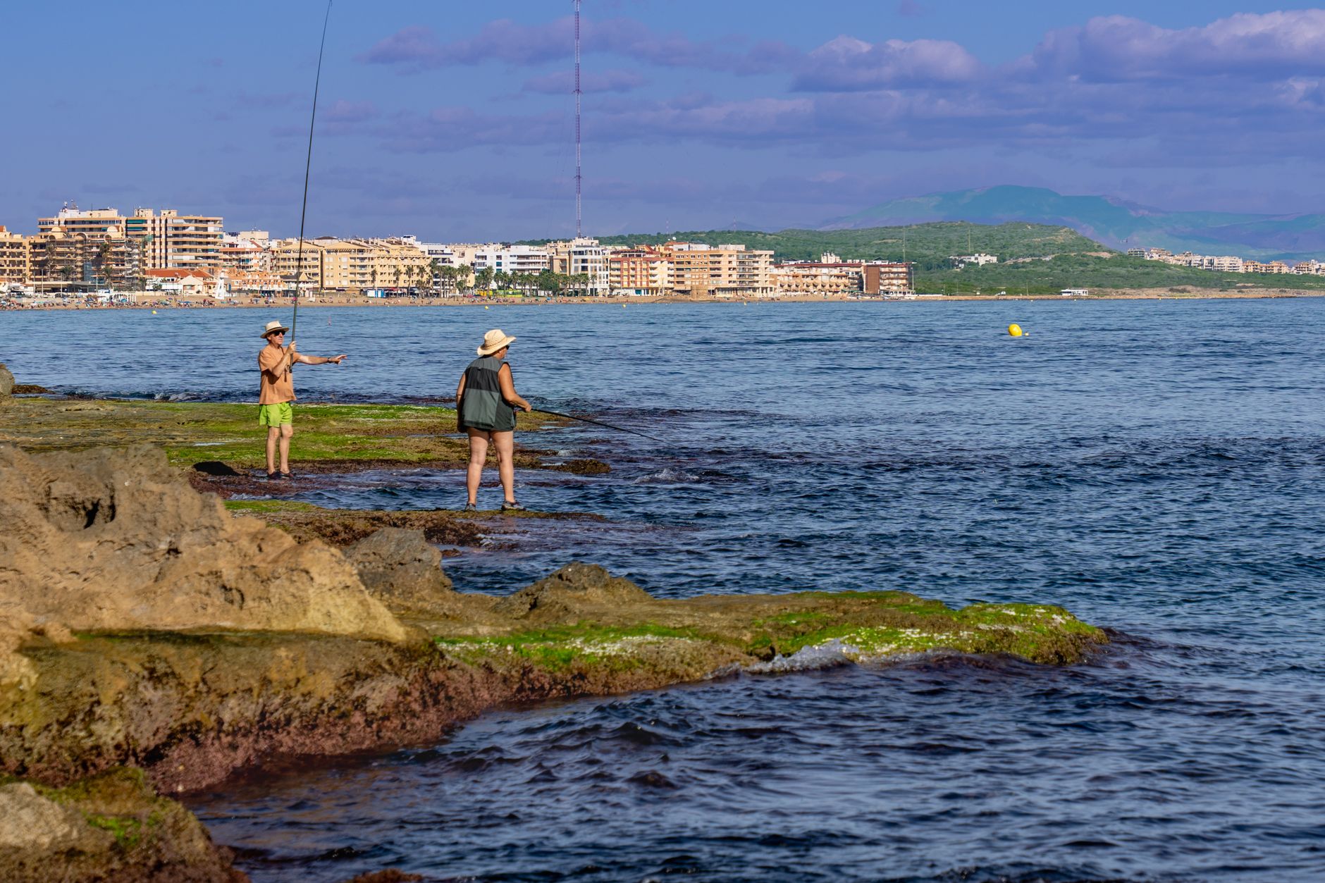 Two people fish on a rocky shoreline with Torrevieja cityscape in the background.